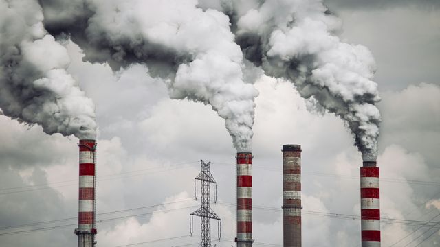 Gray smoke billowing from a number of red and white smokestacks, against a gray cloudy sky. 