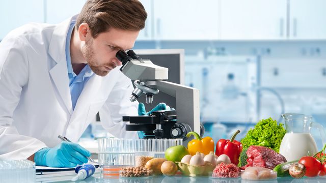 Male scientist looking down a microscope analyzing food samples. A selection of food including fruit, vegetables and meat is laid out on the bench in front of him. 