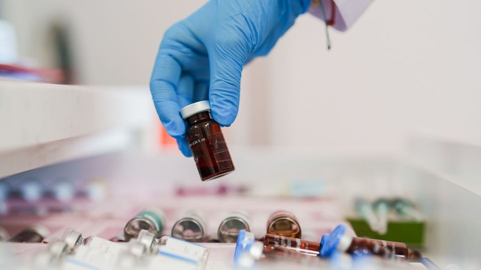 A close-up of a gloved hand holding a small glass vial, with several other vials scattered in the background. The vials are stored in a drawer or tray, suggesting a laboratory or medical setting.